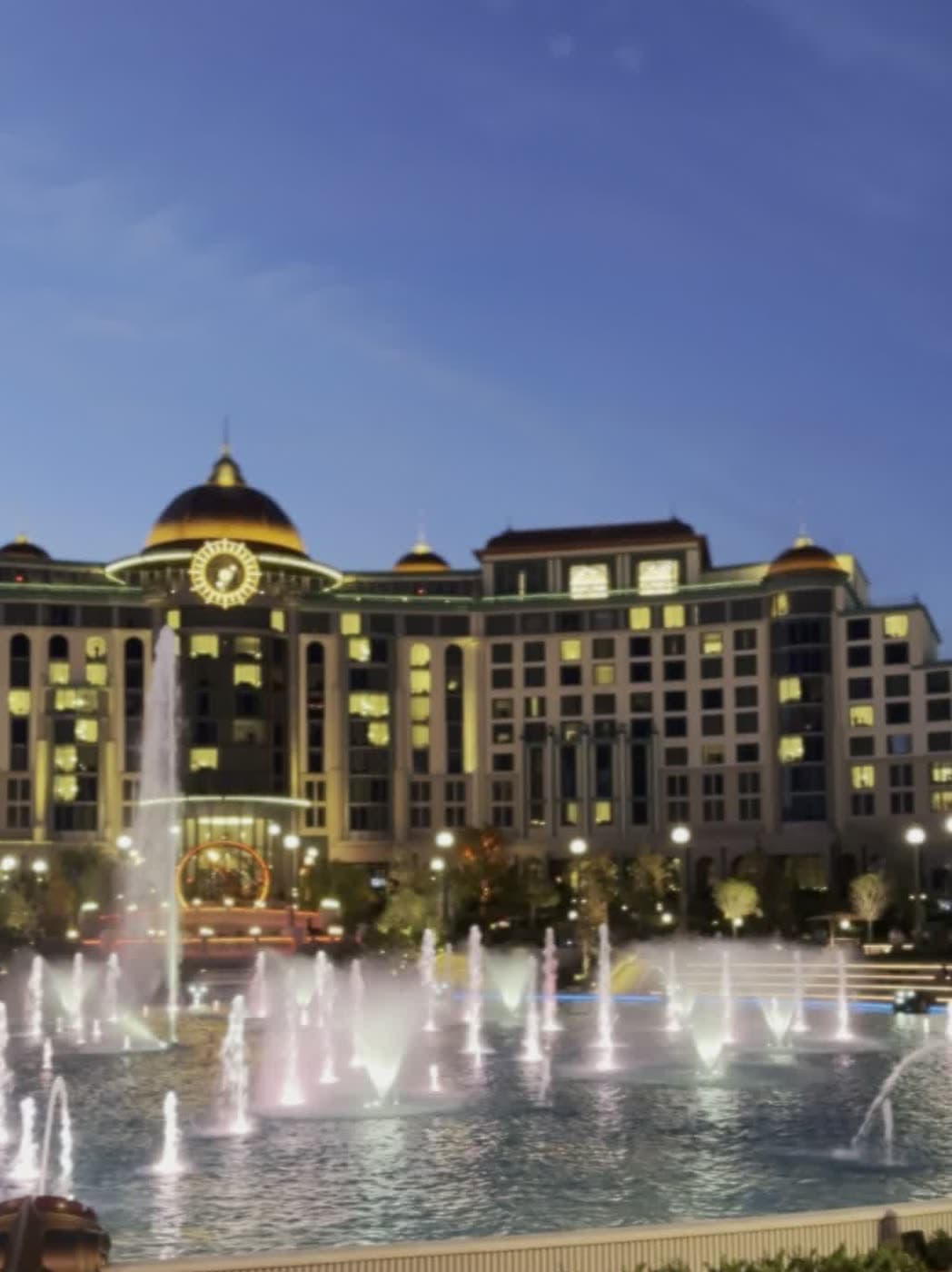 A grand domed hotel lit up at dusk behind dancing lake fountains